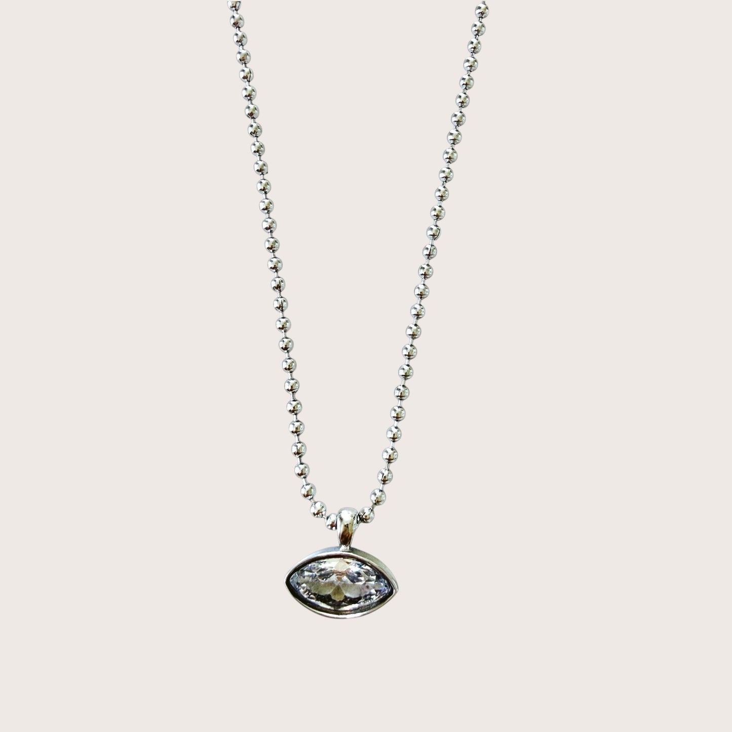A silver beaded chain necklace with an oval bezel-set clear gemstone pendant, shown against a solid light beige background.