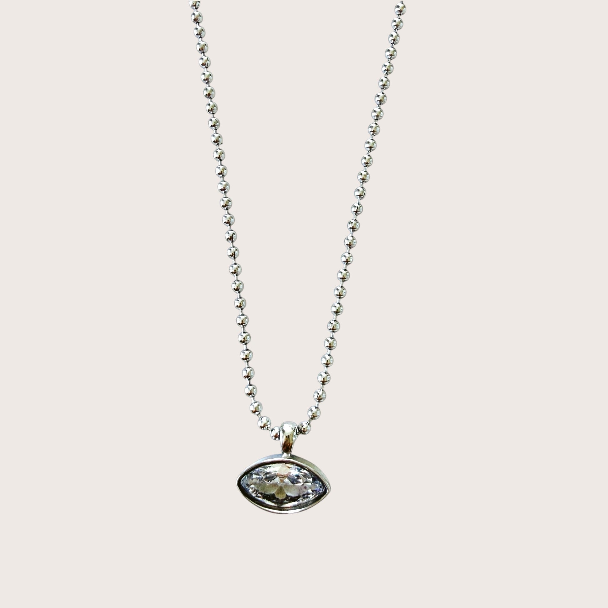 A silver beaded chain necklace with an oval bezel-set clear gemstone pendant, shown against a solid light beige background.