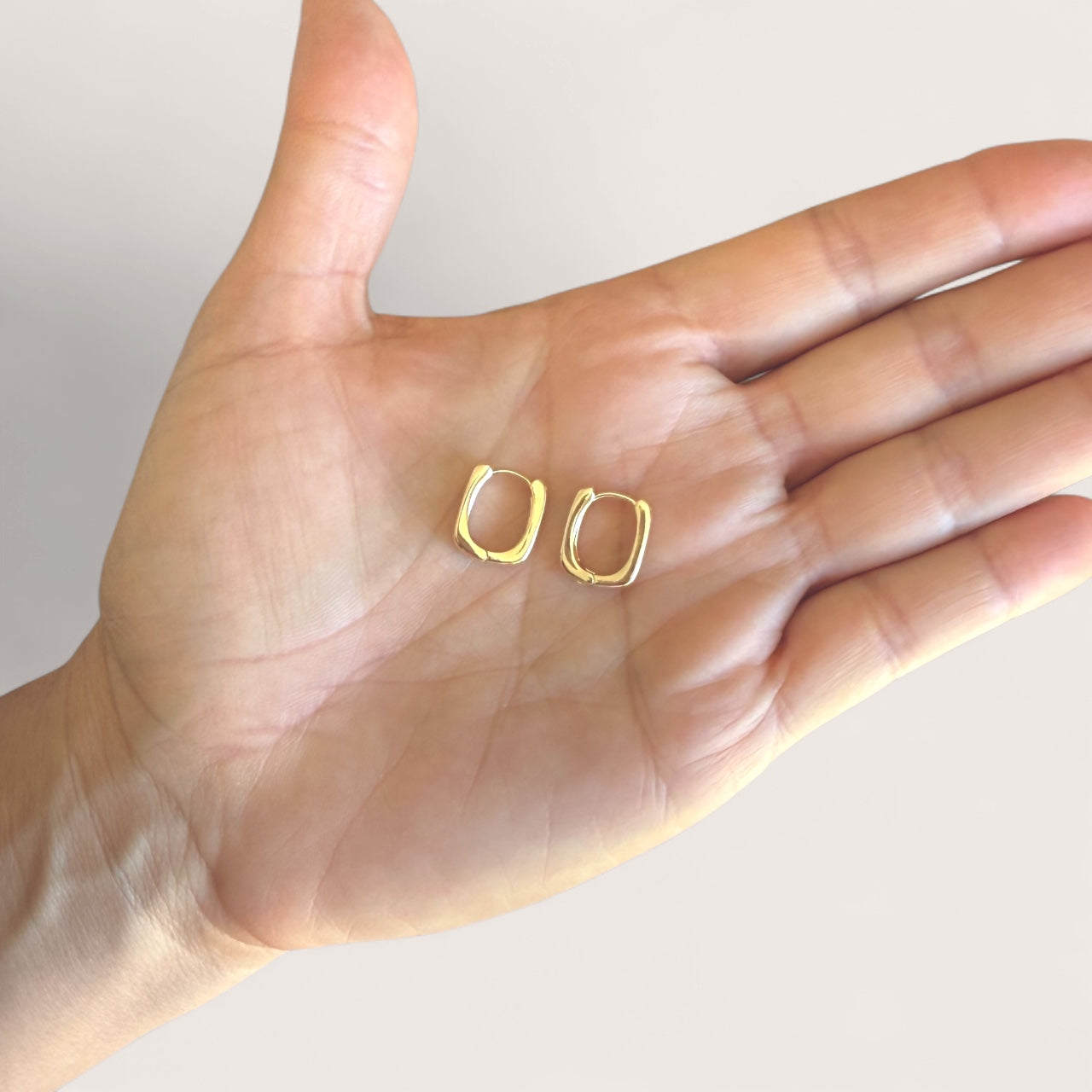 Gold hoop earrings held in the palm of a hand against a neutral background