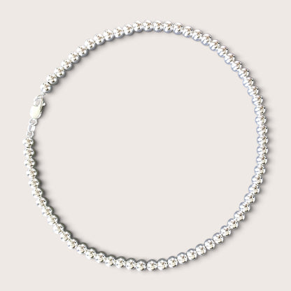 Flat lay of a solid 925 sterling silver 6mm round bead necklace over a black v-neck top against a warm minimalist studio background. The beads have a mirror-bright shine and are precisely scaled to 6mm.