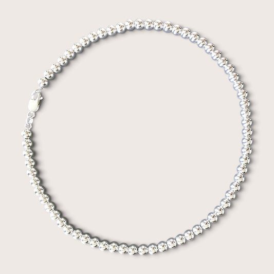 Flat lay of a solid 925 sterling silver 6mm round bead necklace over a black v-neck top against a warm minimalist studio background. The beads have a mirror-bright shine and are precisely scaled to 6mm.