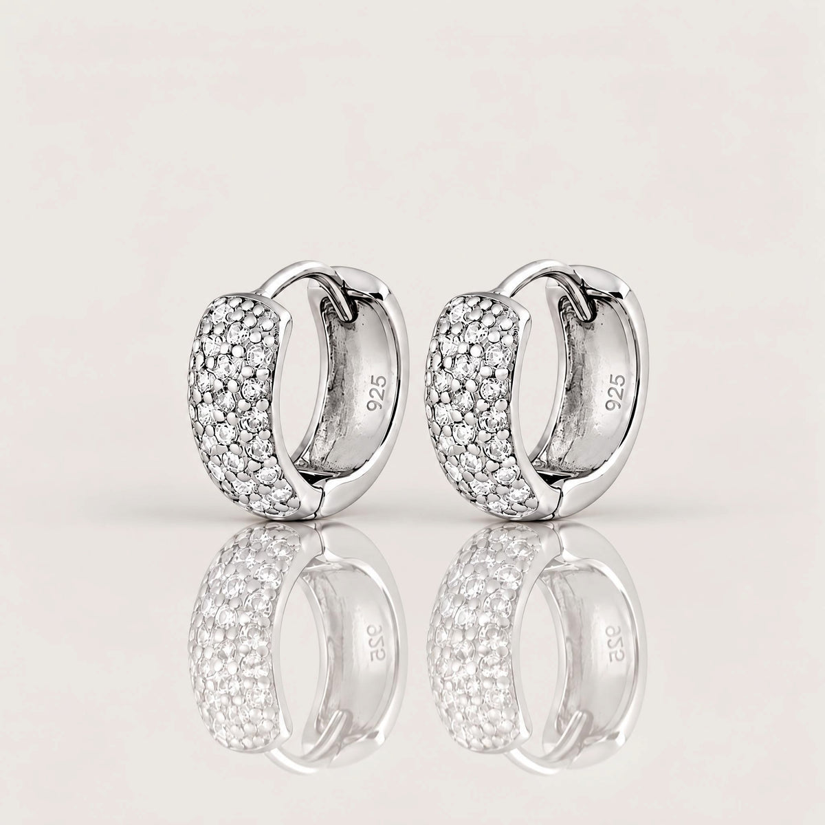 A pair of silver huggie hoop earrings with pavé-set clear stones on the front, shown on a light beige reflective surface. The inside of the earrings shows a 925 sterling silver hallmark.