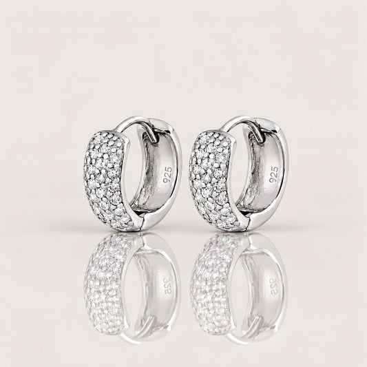 A pair of silver huggie hoop earrings with pavé-set clear stones on the front, shown on a light beige reflective surface. The inside of the earrings shows a 925 sterling silver hallmark.