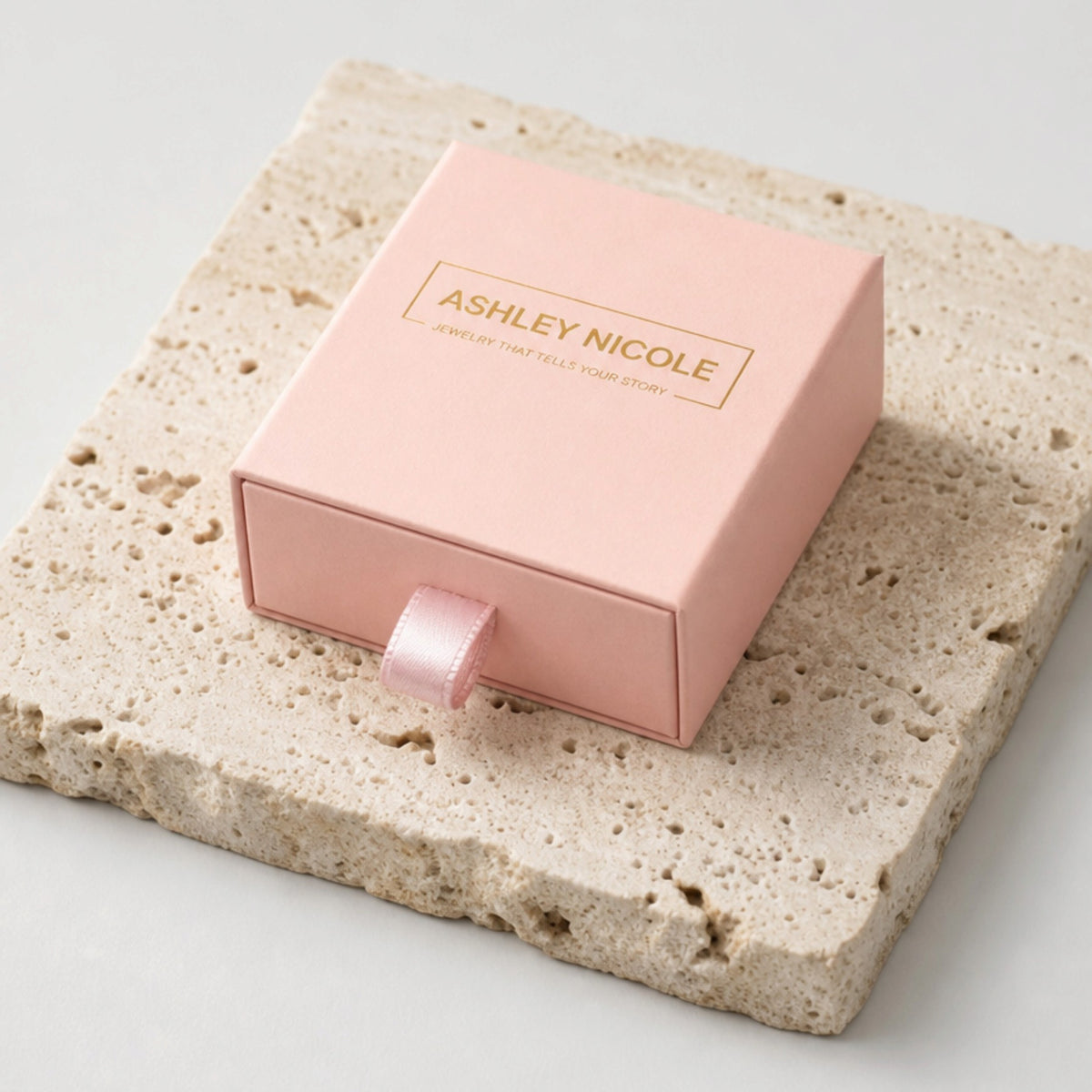 Pink Ashley Nicole Jewelry drawer-style gift box with a ribbon pull on a stone surface