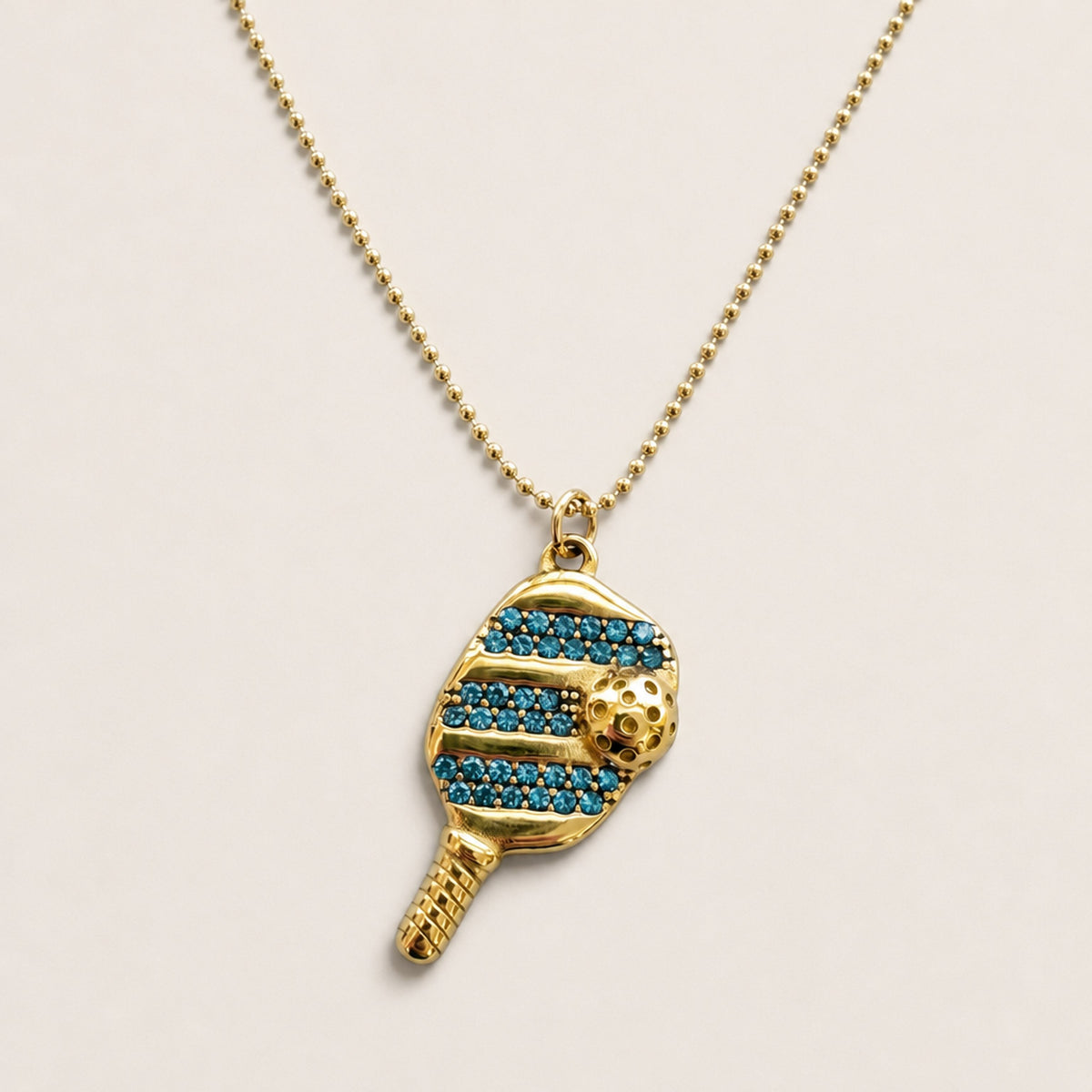 Blue cubic zirconia pickleball paddle-and-ball pendant on a fine gold ball chain, shown from the front.