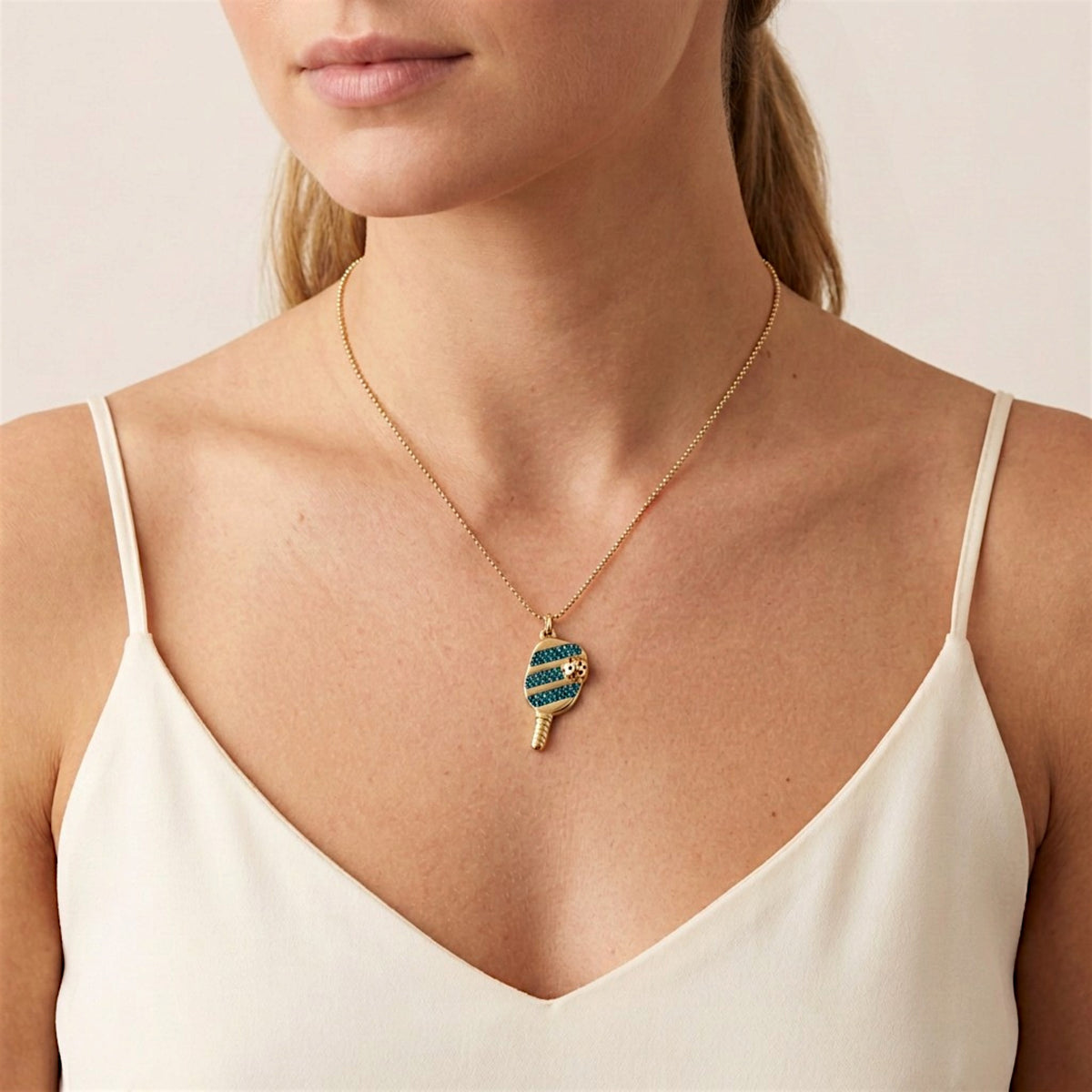 Woman wearing a blue cubic zirconia pickleball paddle pendant on a fine gold chain above an ivory neckline.