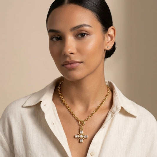 A woman wearing a gold cross pendant necklace with clear crystals, styled with a white linen shirt against a warm beige background.