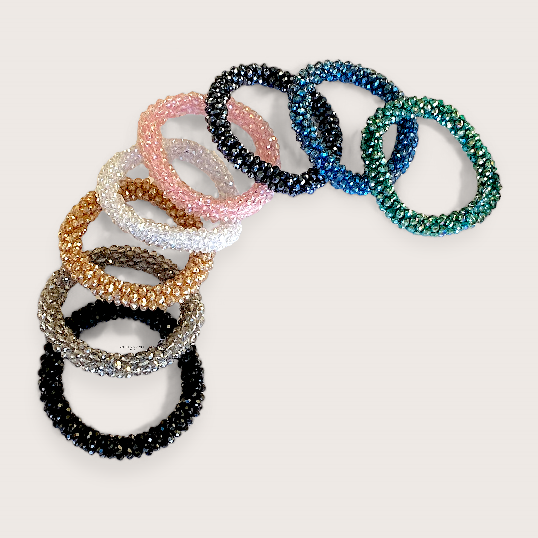 A collection of crystal bead elastic hair ties laid flat on a clean background showing the multi-faceted in black, silver, gold, teal blue, white, and pink, glass beads and woven stretch band by Ashley Nicole Jewelry. 