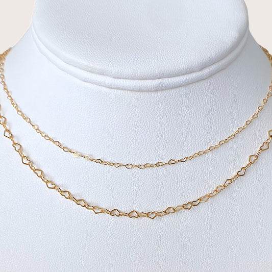 Two delicate gold chain necklaces displayed on a white textured jewelry bust. The top chain is a fine link style, while the bottom chain features repeating open heart-shaped links.