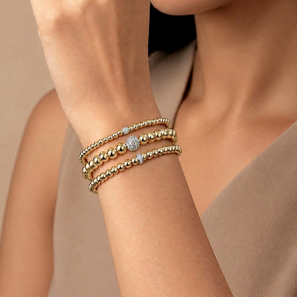 Three gold beaded bracelets with diamond accents worn together on a raised wrist beside a taupe top