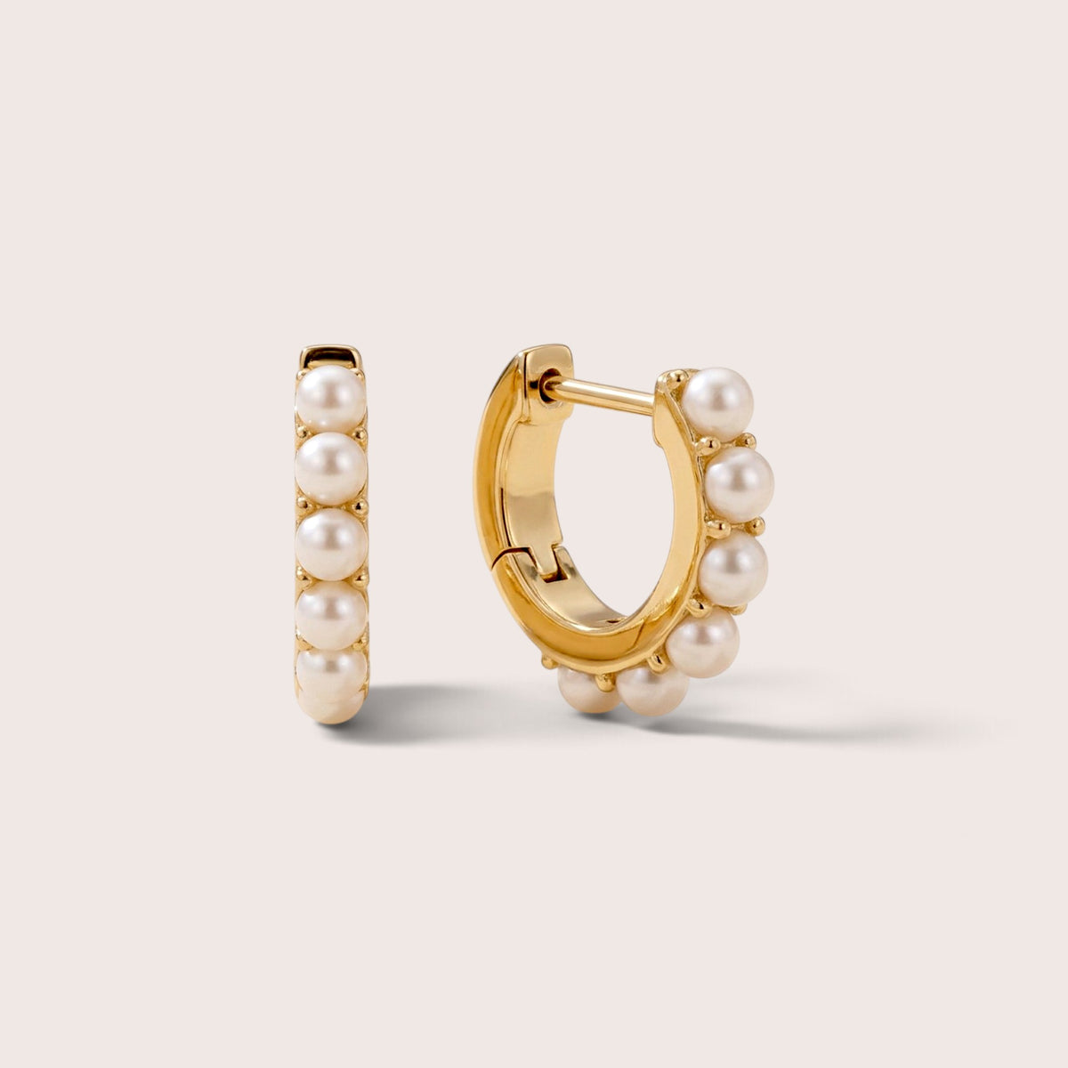 Gold huggie hoops with round white freshwater pearls, shown from the front and at an angle on a warm neutral background.