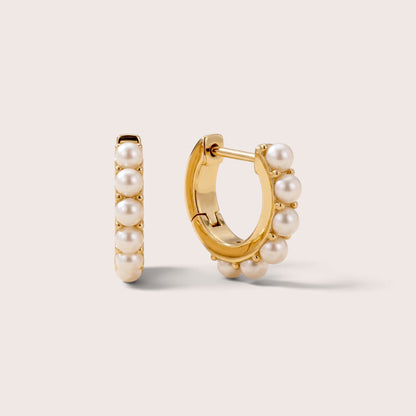Gold huggie hoops with round white freshwater pearls, shown from the front and at an angle on a warm neutral background.