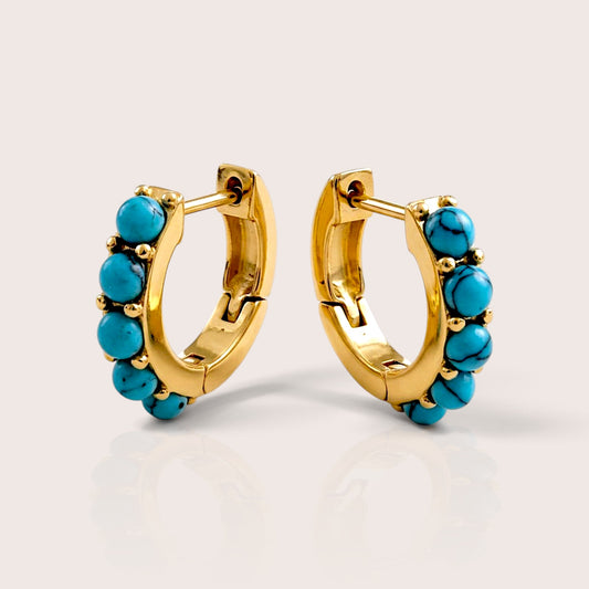 Pair of gold huggie hoops with round turquoise stones and dark veining, shown at angled views on a warm neutral background.