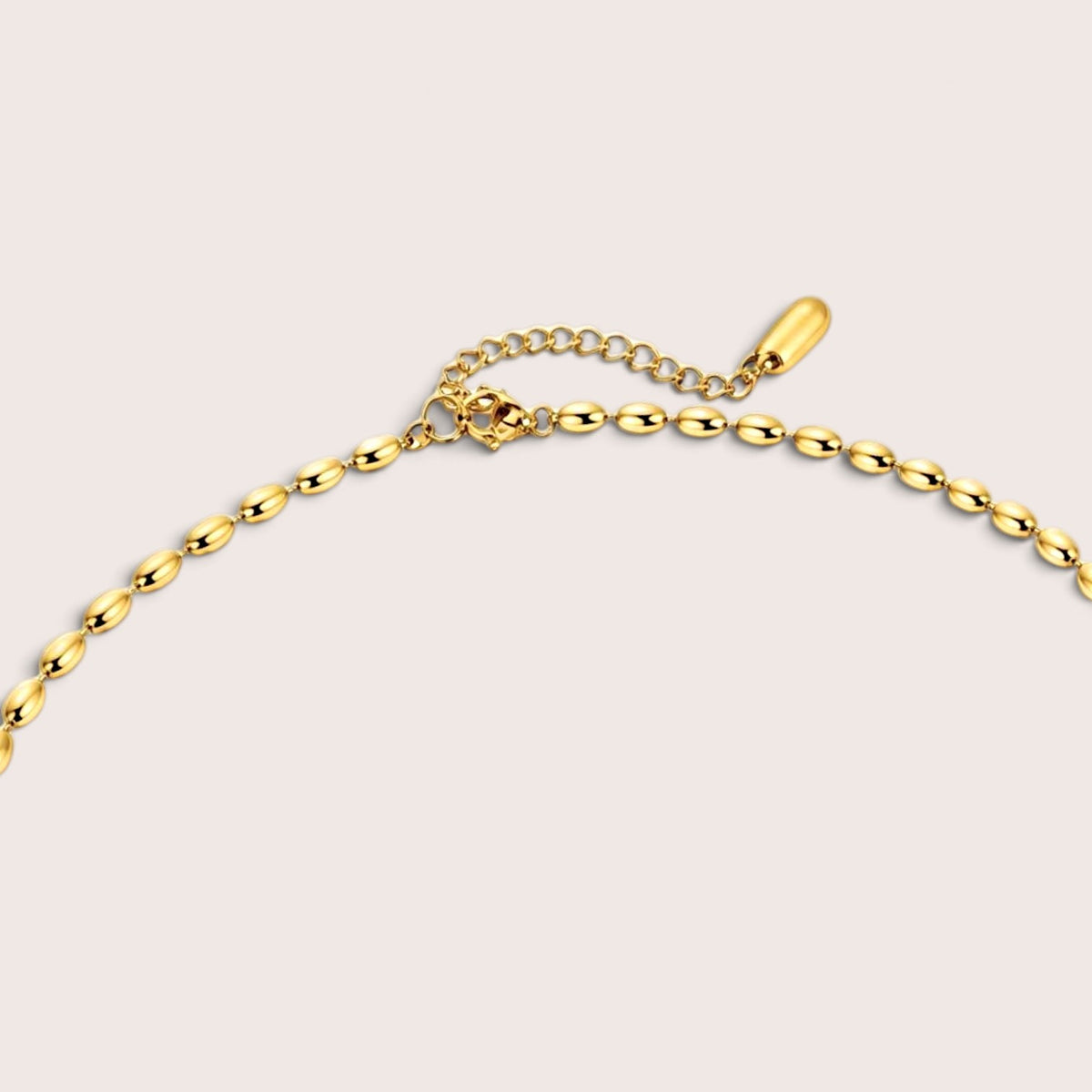 Close-up of a gold chain necklace with elongated oval links, an extension chain, and a small polished gold tag, shown on a light beige background.