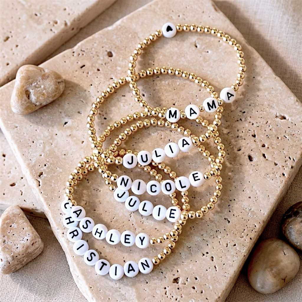 Five personalized beaded bracelets with gold-toned round beads and white letter tiles spelling names or words, arranged on beige stone tiles with small stones around them.
