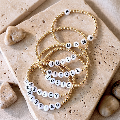 Five personalized beaded bracelets with gold-toned round beads and white letter tiles spelling names or words, arranged on beige stone tiles with small stones around them.