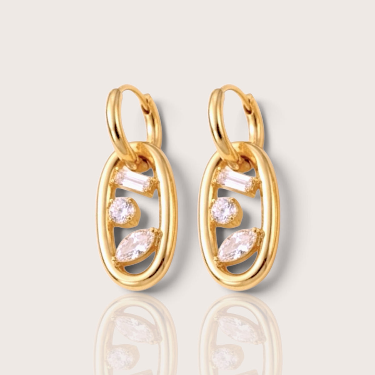 A pair of gold huggie hoop earrings with oval links and clear marquise and baguette stones, displayed on a light beige background.