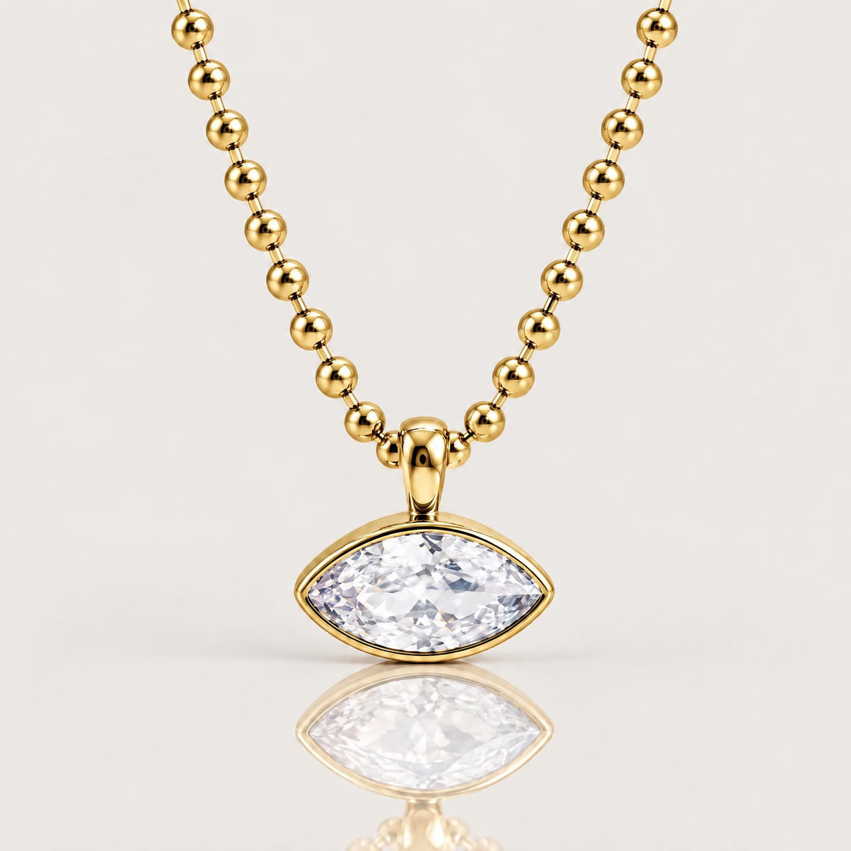 A gold beaded necklace with a marquise-cut clear gemstone pendant in a gold setting, displayed on a clean light gray background with a soft reflection below.