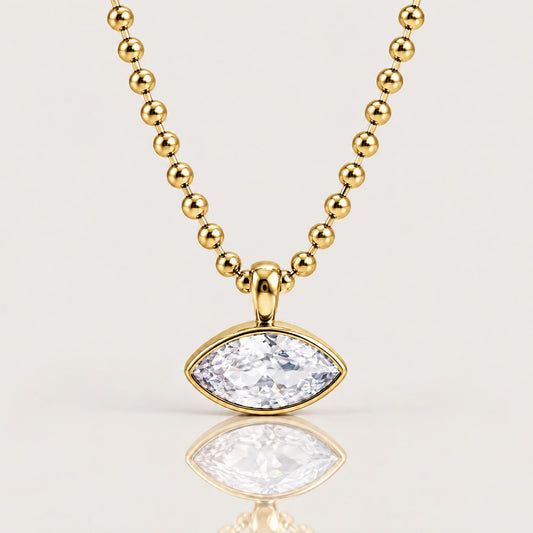 A gold beaded necklace with a marquise-cut clear gemstone pendant in a gold setting, displayed on a clean light gray background with a soft reflection below.