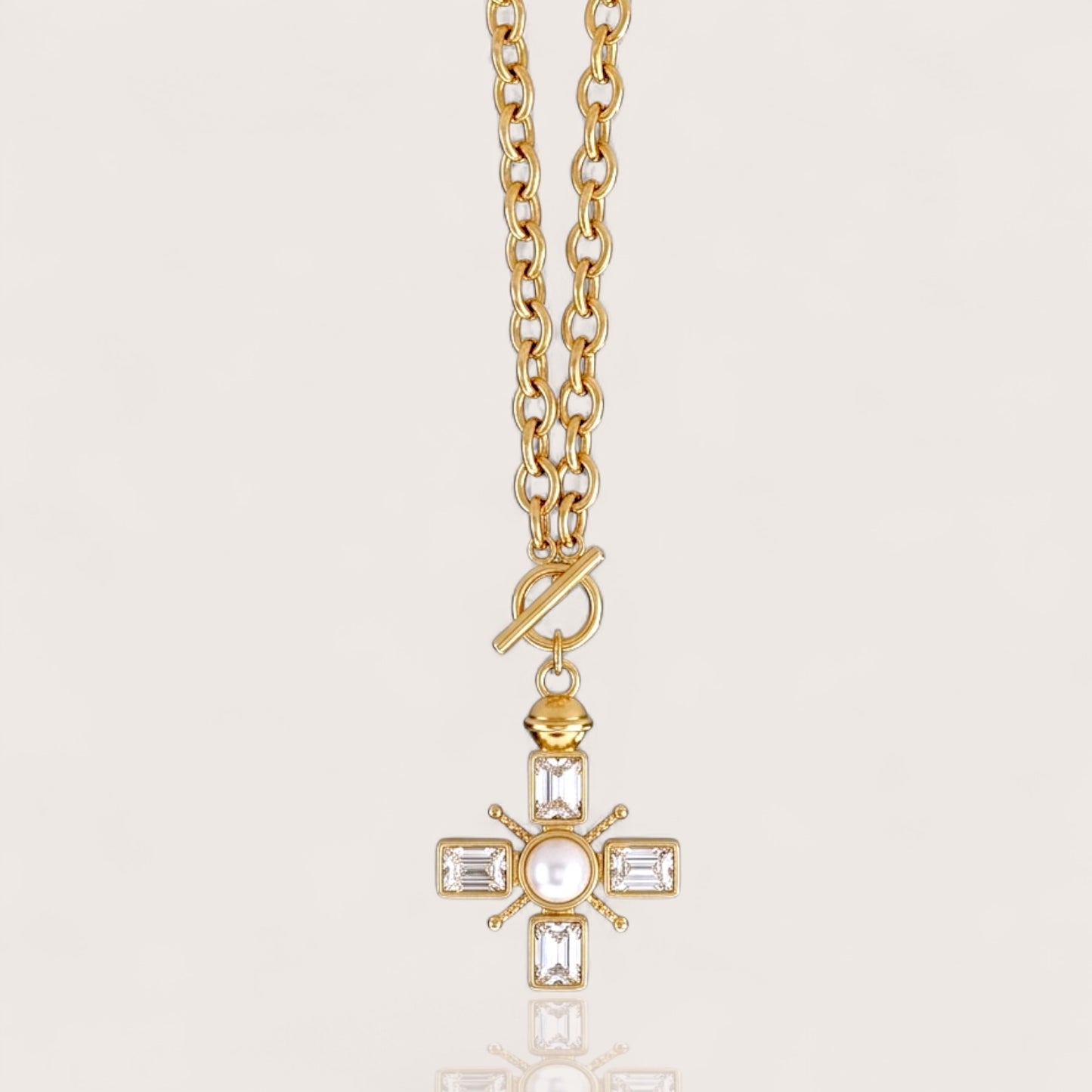 A chunky gold chain necklace with a toggle clasp and a cross-shaped pendant featuring clear baguette and round gemstones, shown against a light beige background.