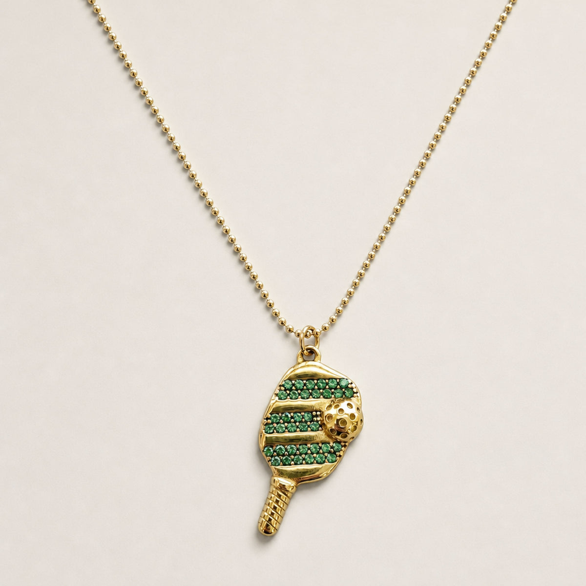 Green cubic zirconia pickleball paddle-and-ball pendant hanging from a fine gold ball chain on a warm neutral background.