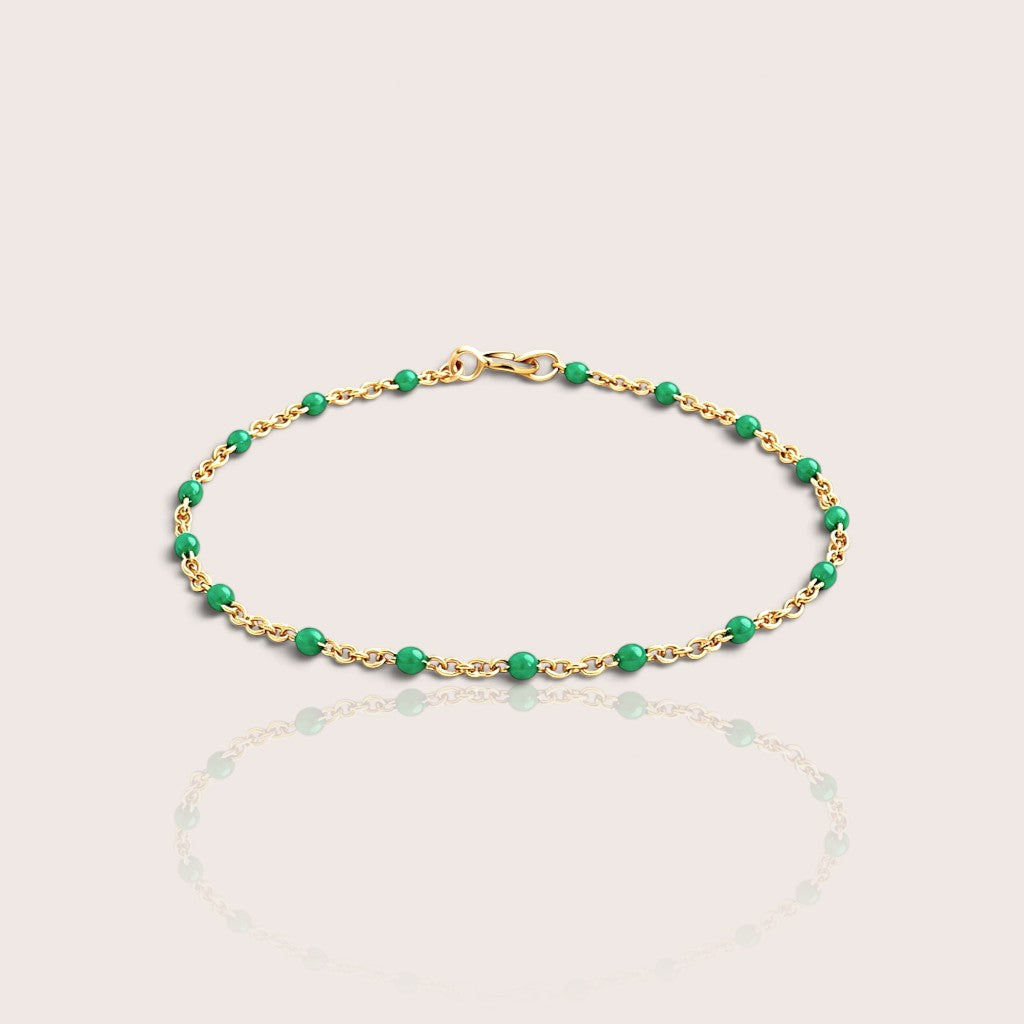 A delicate gold chain bracelet with evenly spaced, polished green beads, displayed on a light beige background with a soft reflection below.