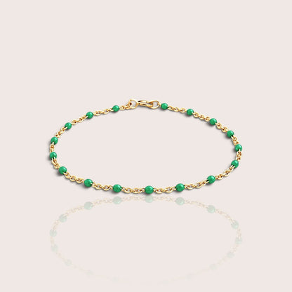 A delicate gold chain bracelet with evenly spaced, polished green beads, displayed on a light beige background with a soft reflection below.