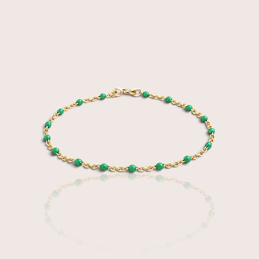 A delicate gold chain bracelet with evenly spaced, polished green beads, displayed on a light beige background with a soft reflection below.