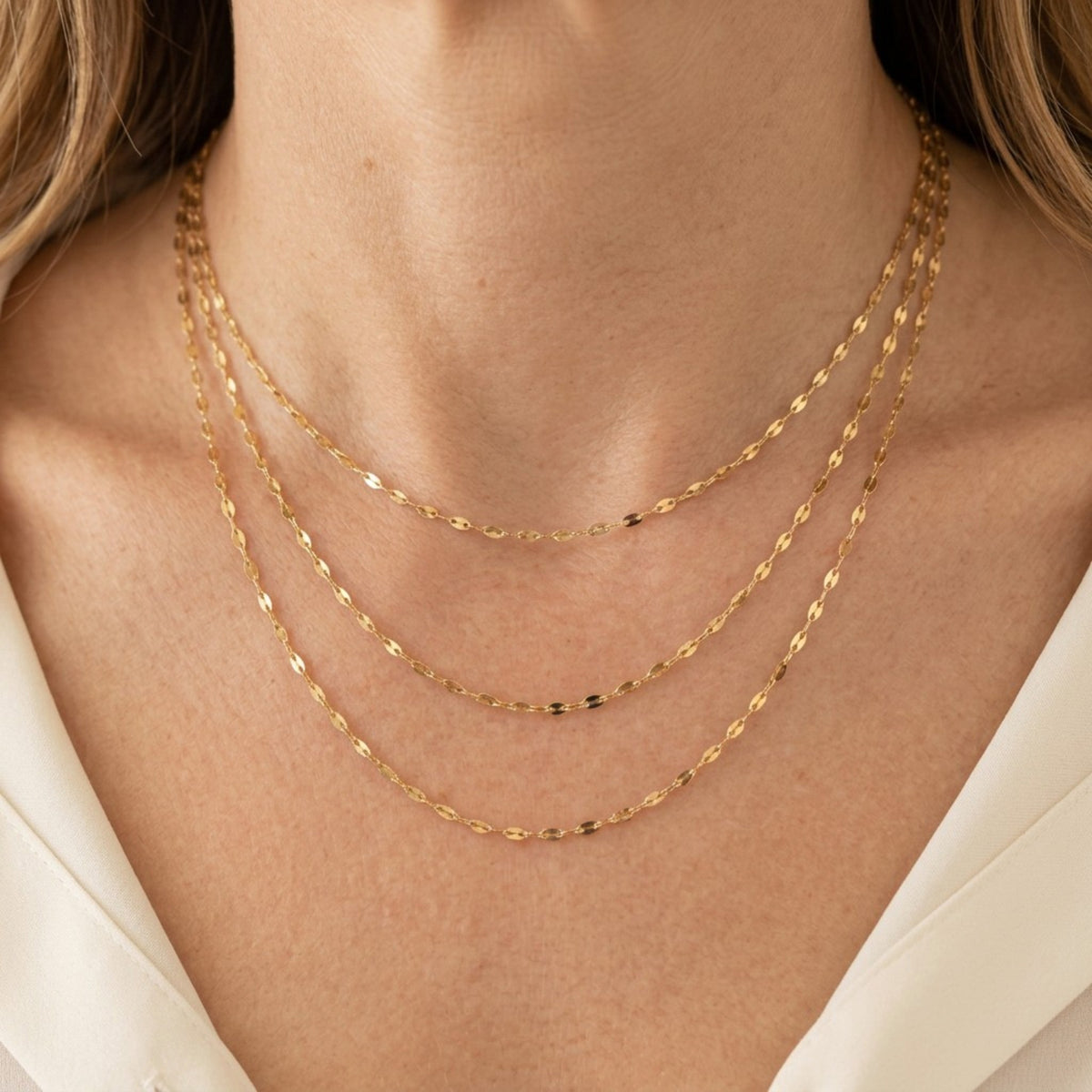 Three gold-filled mirror Valentino chain necklaces worn at different lengths above an ivory blouse