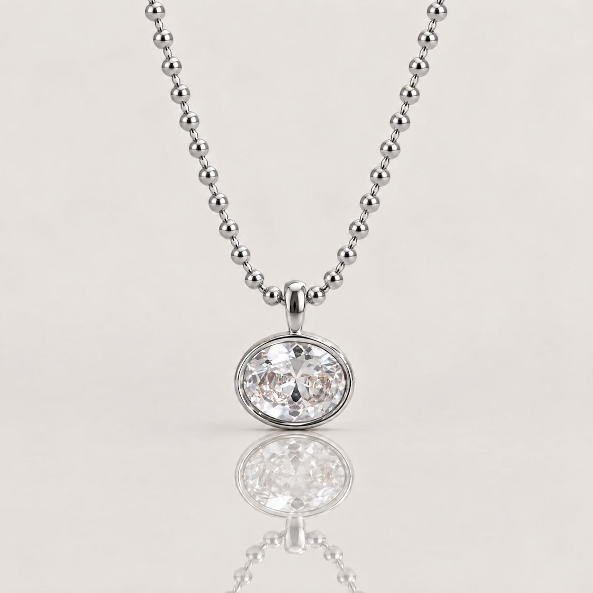 A silver beaded ball chain necklace with a round bezel-set clear gemstone pendant, shown on a light beige background with a soft reflection below.
