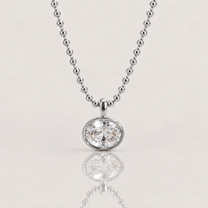 A silver beaded ball chain necklace with a round bezel-set clear gemstone pendant, shown on a light beige background with a soft reflection below.