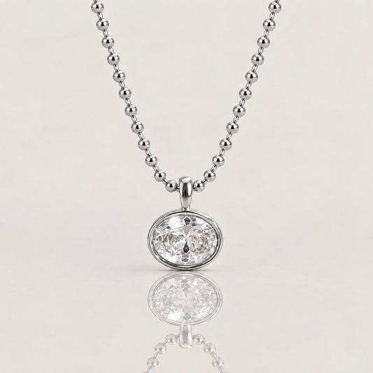 A silver beaded ball chain necklace with a round bezel-set clear gemstone pendant, shown on a light beige background with a soft reflection below.