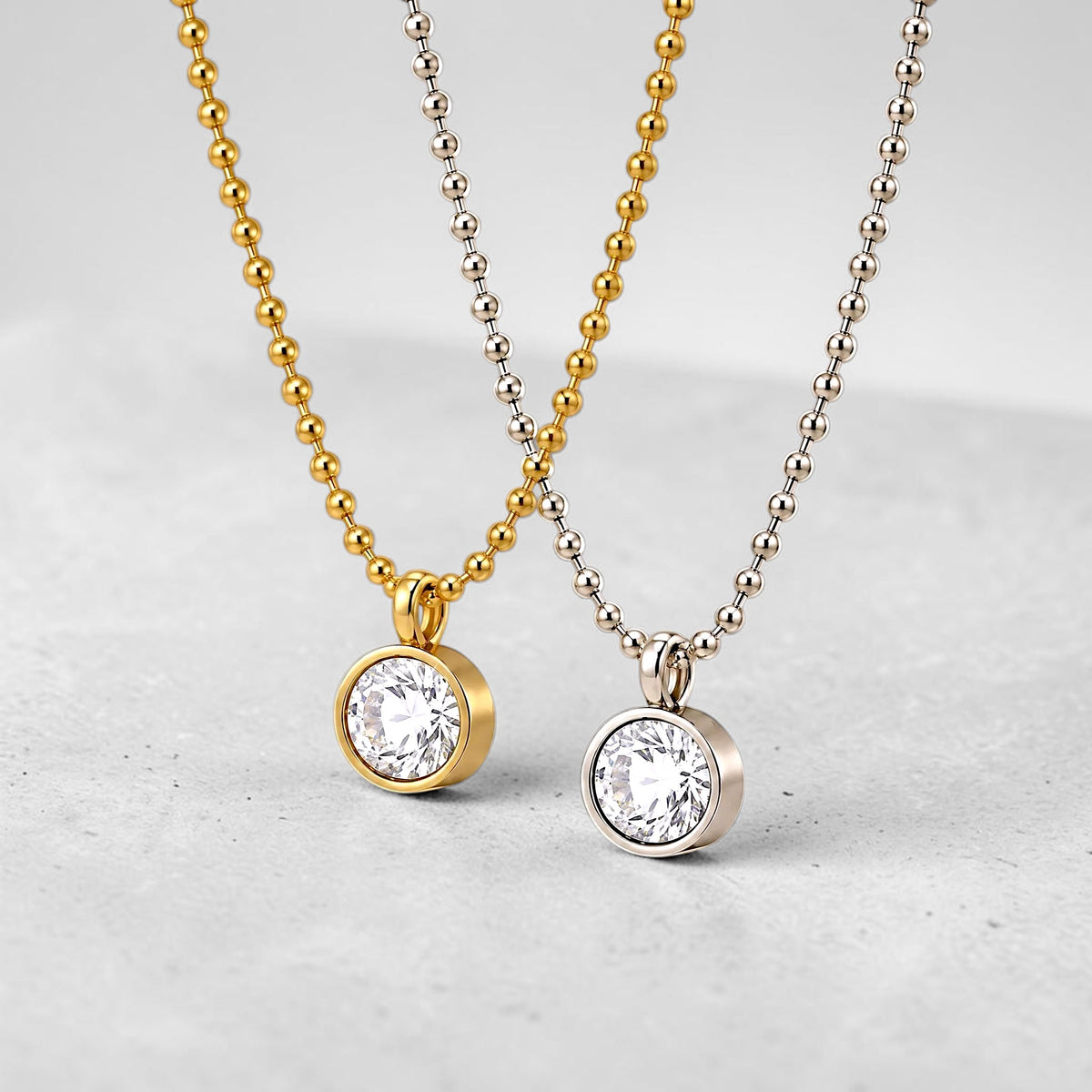 Two beaded ball-chain necklaces with round bezel-set clear gemstones, one in gold-toned metal and one in silver-toned metal, displayed on a light gray surface.