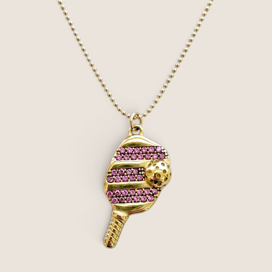 Gold pickleball paddle pendant with pink pavé cubic zirconia and a raised gold ball, 
shown at an angle on a ball chain.