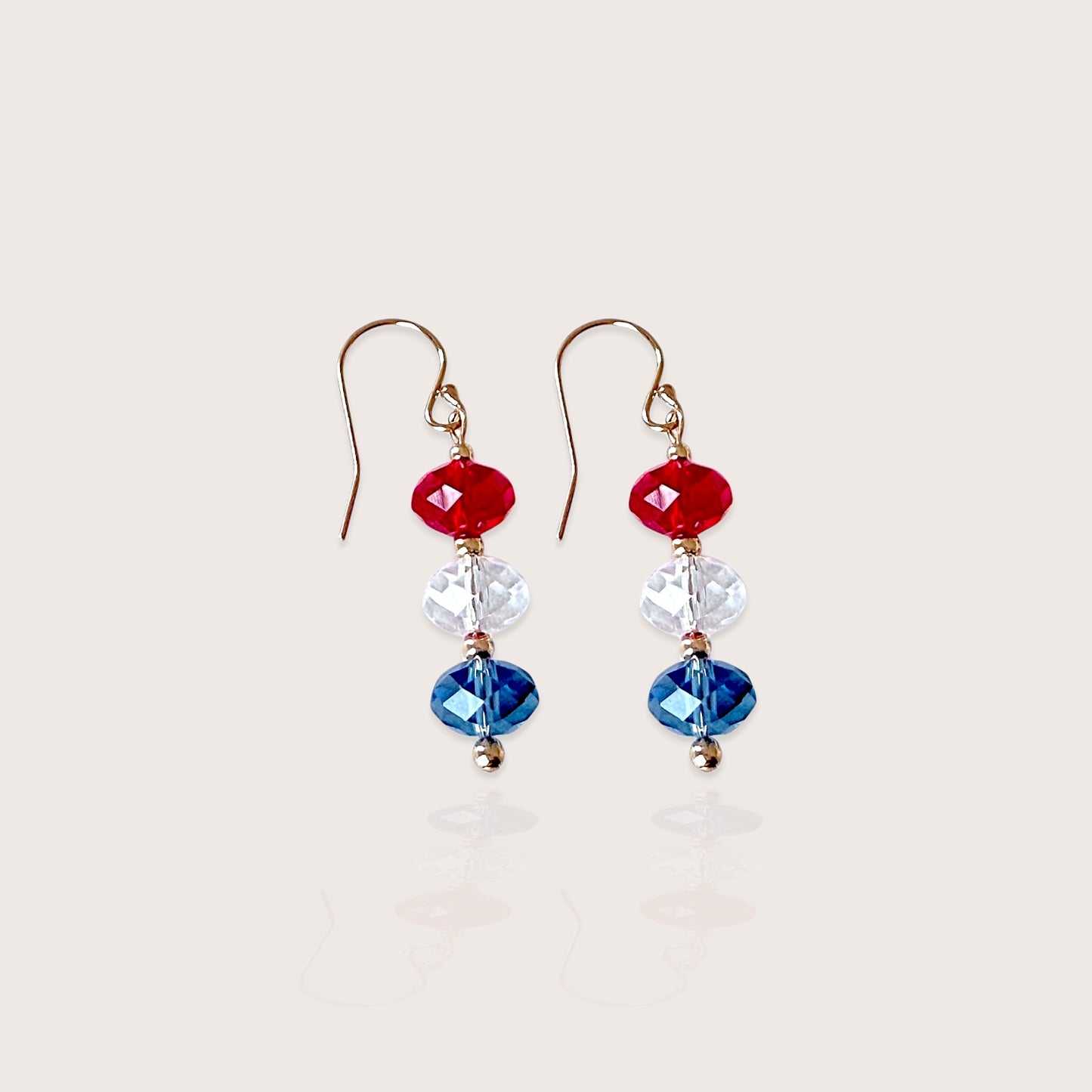 A close-up photo of handcrafted 14k gold-filled patriotic dangle drop earrings featuring a vertically stacked red white and blue Swarovski crystal bead design on a French wire hook by Ashley Nicole Jewelry.