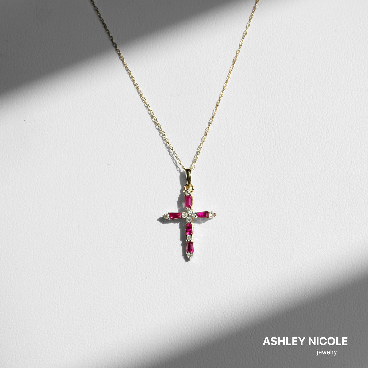 Ruby and diamond cross necklace on a light surface in directional sunlight, with the fine gold chain visible