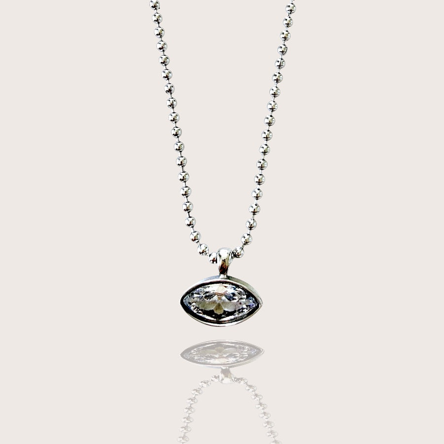 A silver beaded chain necklace with an oval bezel pendant containing a clear faceted gemstone, shown against a light beige background with a soft reflection below.