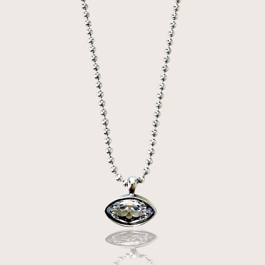 A silver beaded chain necklace with an oval bezel pendant containing a clear faceted gemstone, shown against a light beige background with a soft reflection below.