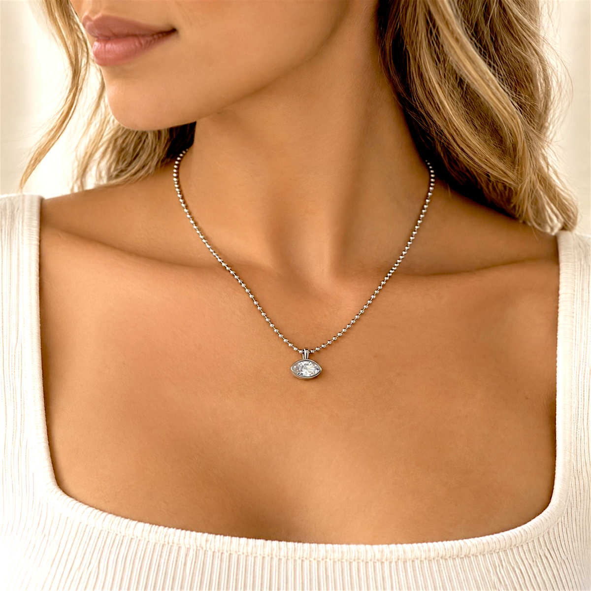 A close-up lifestyle shot of a woman wearing a delicate silver chain necklace with a small silver eye pendant, paired with a white scoop-neck t-shirt.