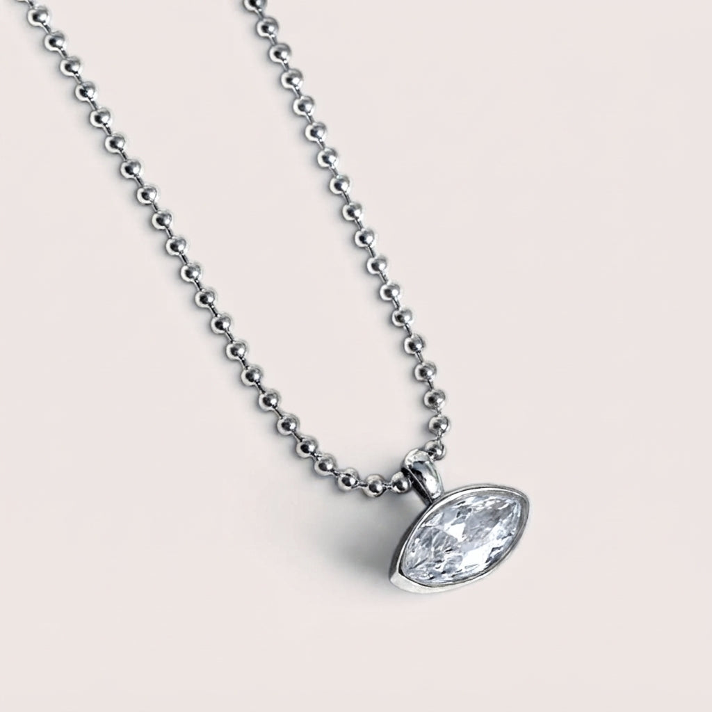 A silver beaded necklace with an oval bezel-set clear gemstone pendant, shown against a solid light beige background.