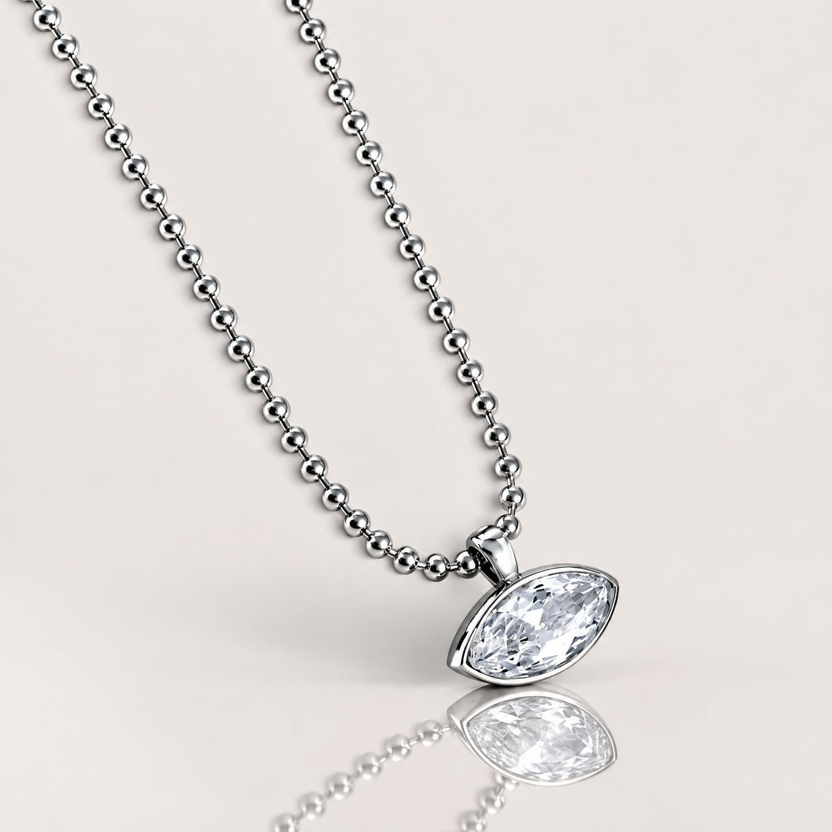 A silver beaded necklace with an oval bezel-set clear gemstone pendant, shown against a solid light beige background.