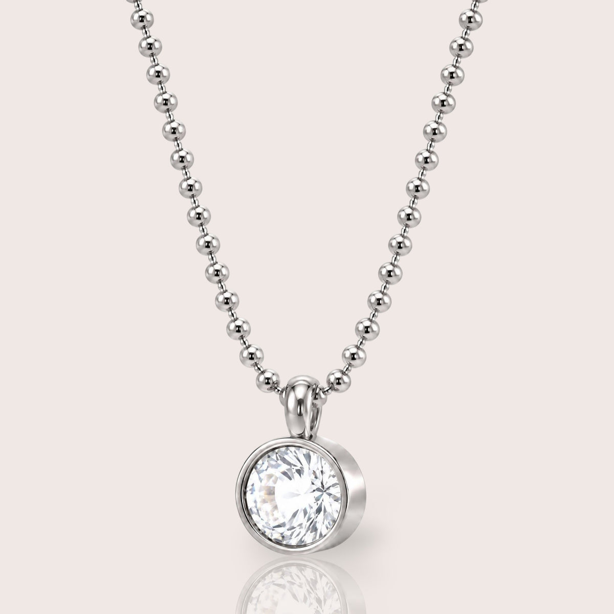 A silver beaded ball chain necklace with a round bezel-set clear gemstone pendant, shown on a light beige background with a soft reflection below.