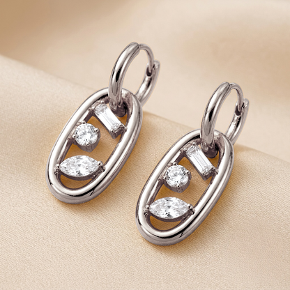 A pair of silver-toned drop earrings with marquise-cut and baguette-cut clear stones, displayed on a soft beige fabric background.