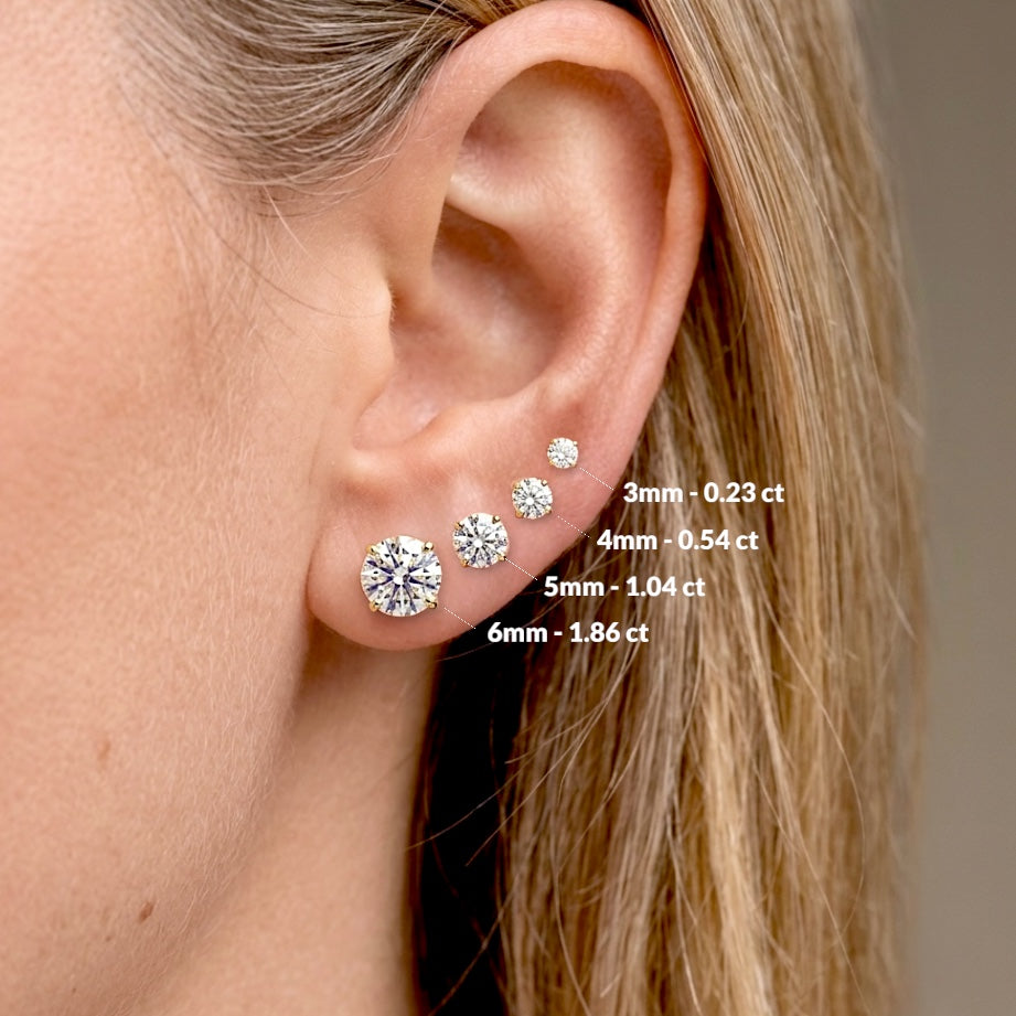 Close-up of an ear with diamond earrings and size labels. Side view of real 14K solid gold round diamond studs in premium VS-VVS diamond clarity.Model wearing earrings for size visual - from small to large 0.23 ctw (3mm), 0.54 ctw (4mm), 1.04 ctw (5mm), 1.86 ctw (6mm)