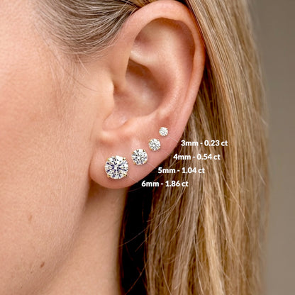 Close-up of an ear with diamond earrings and size labels. Side view of real 14K solid gold round diamond studs in premium VS-VVS diamond clarity.Model wearing earrings for size visual - from small to large 0.23 ctw (3mm), 0.54 ctw (4mm), 1.04 ctw (5mm), 1.86 ctw (6mm)