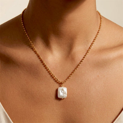 Woman wearing a gold ball chain necklace with a white baroque pearl pendant above an ivory neckline.
