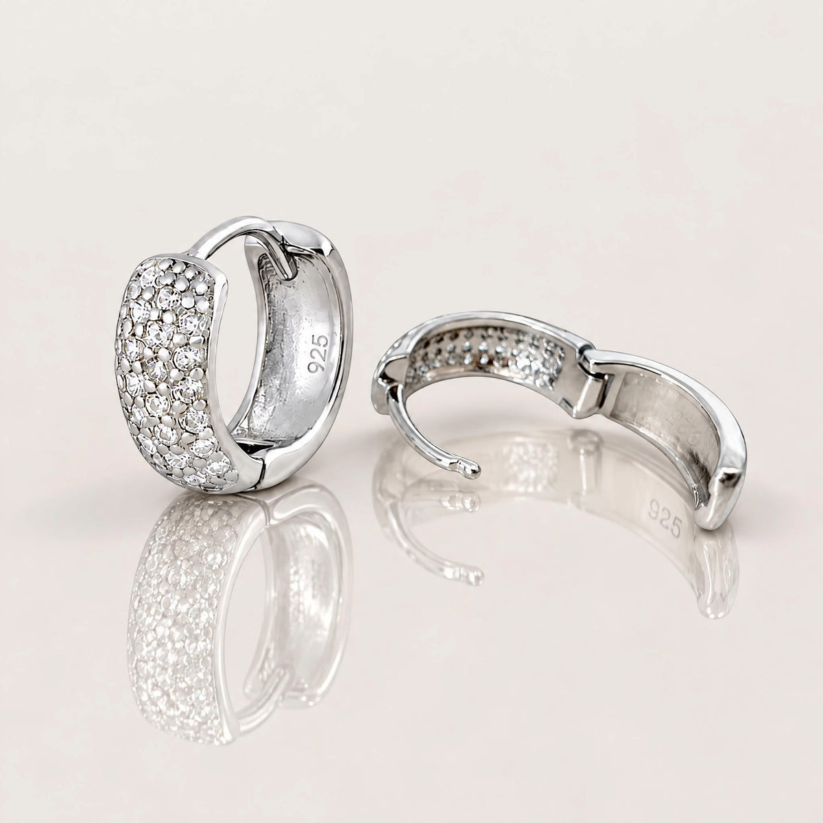 A pair of silver huggie hoop earrings paved with small clear stones, shown on a light beige reflective surface. One earring is facing forward, showing a '925' sterling silver hallmark, and the other is turned to show the back clasp mechanism.