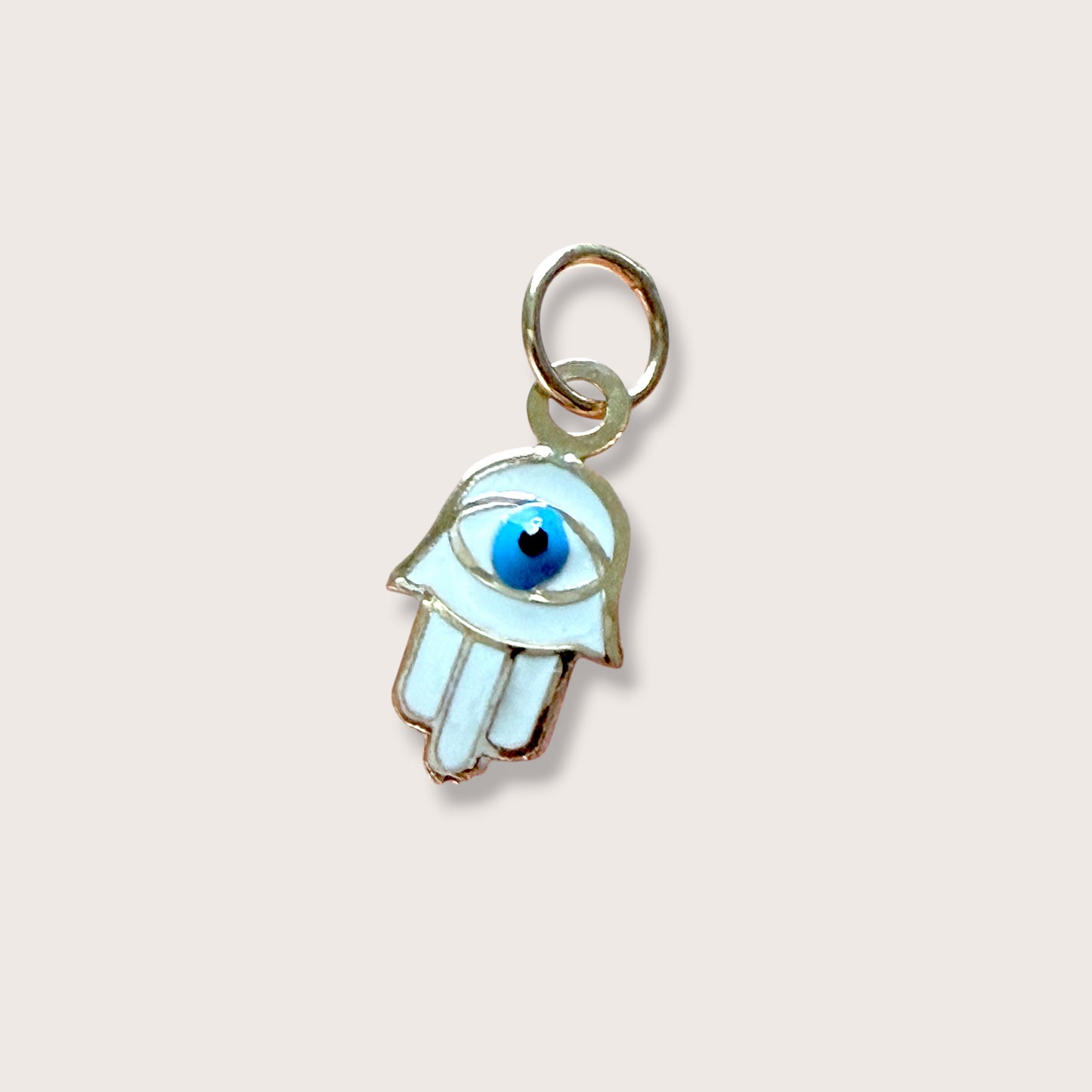 A small Hamsa hand charm with a white enamel body, gold-tone metal outline, and a blue-and-black evil eye detail, shown on a plain light beige background.