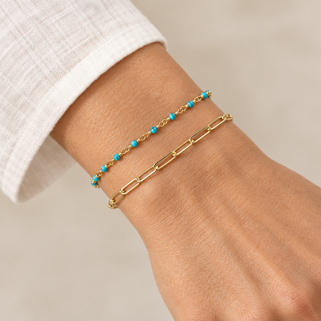 Turquoise bead bracelet layered with a gold-tone paperclip bracelet on a wrist beside a white sleeve.