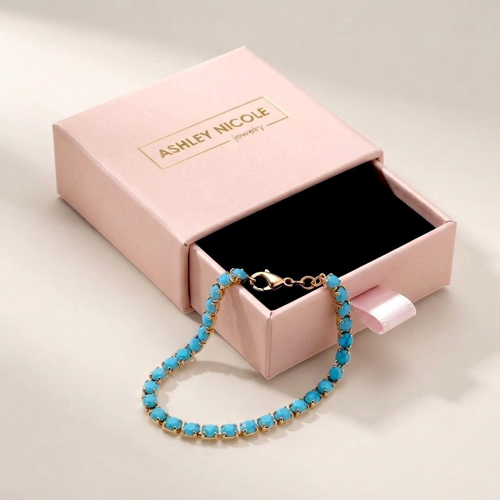 the gold and turquoise tennis bracelet resting next to its open, branded pink jewelry gift box from Ashley Nicole Jewelry.